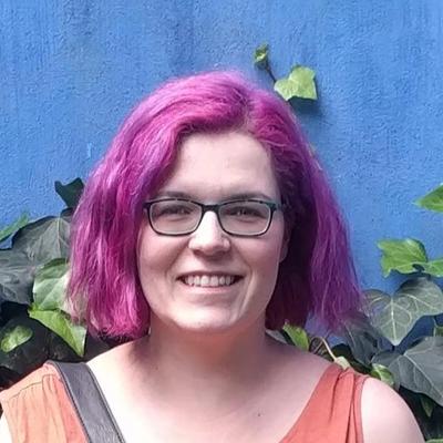 Portrait of a woman with pink short hair, wearing glasses and an orange sleeveless top. Blue background with ivy. 